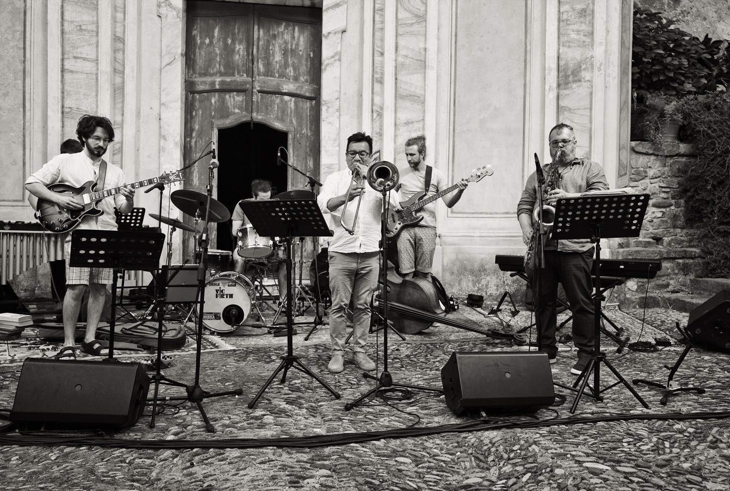 Un gruppo jazz - chitarra, batteria, trombone, basso elettrico e sax - suona davanti a un antico portone di legno, su un selciato di ciottoli, in bianco e nero