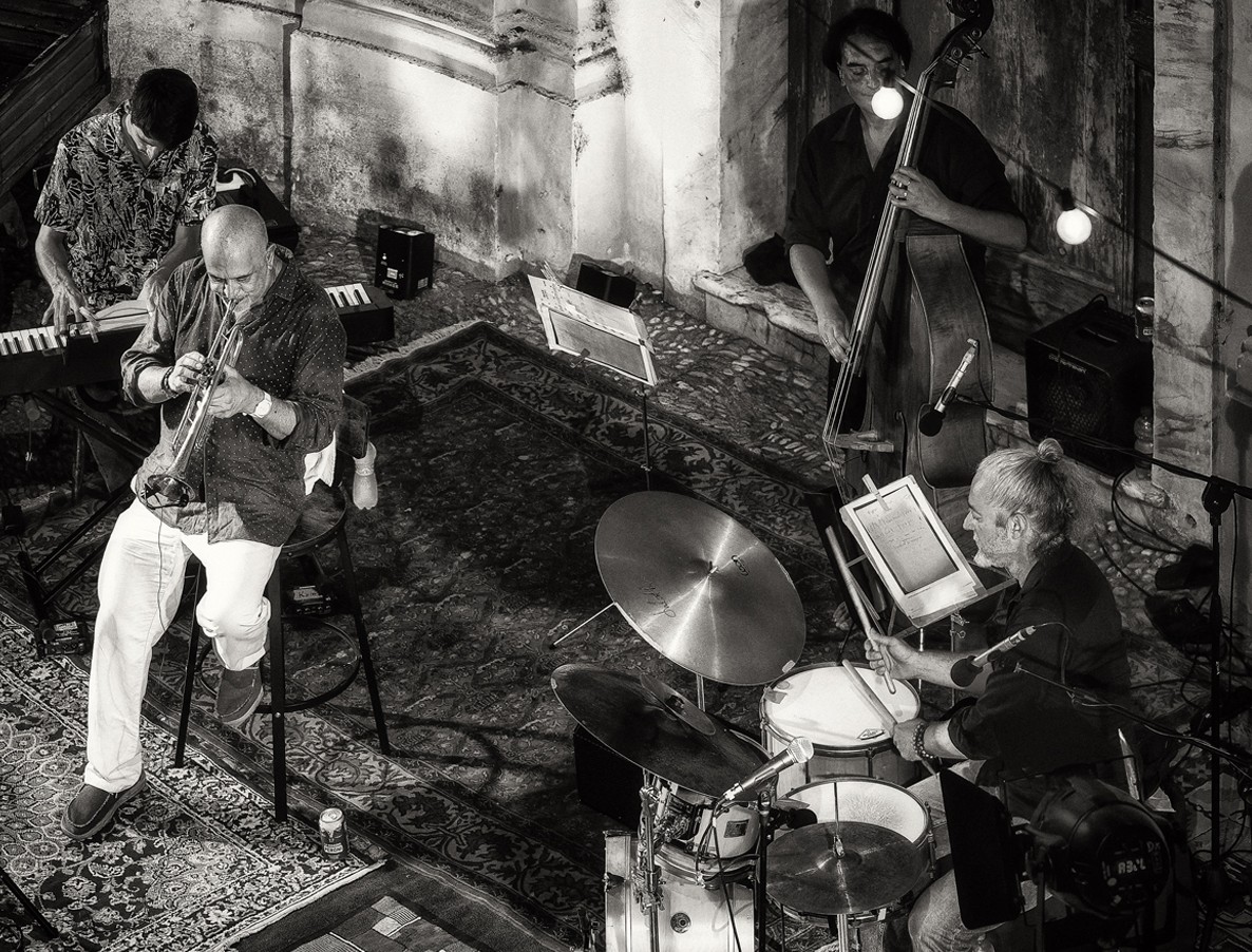 Un quartetto jazz - tromba, tastiera, contrabbasso e batteria - suona in un cortile di pietra, in bianco e nero